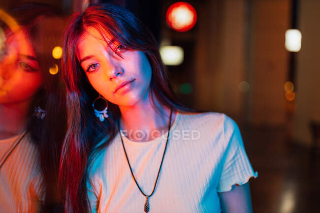 Young gentle female in pendant and earring with long hair looking at camera while leaning on glass wall in evening — Stock Photo