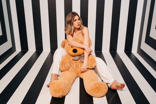 Cheerful barefoot woman in white pants with soft bear sitting on striped floor while looking away — Stock Photo