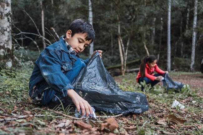Ethnic volunteers with plastic bags picking rubbish from terrain against trees in summer woods in daylight — Stock Photo