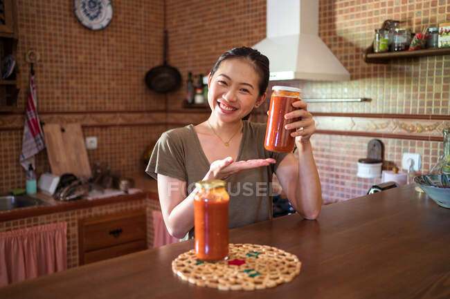 Весела етнічна домогосподарка показує скляні банки з домашнім томатним соусом маринари, сидячи за столом на кухні і дивлячись на камеру — стокове фото