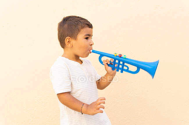 Vista lateral de lindo niño jugando trompeta de juguete azul de pie cerca de la pared naranja claro - foto de stock