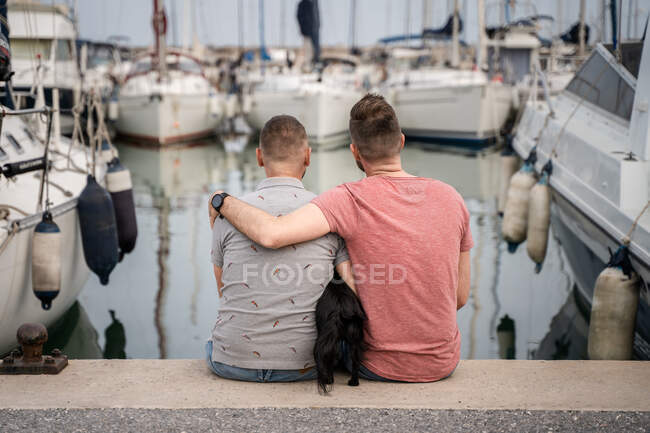 Visão traseira do cão entre homem barbudo alegre abraçando parceiro homossexual anônimo enquanto conversa e se senta no cais no porto — Fotografia de Stock