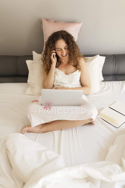 De cima mulher de negócios com cabelo encaracolado sentado na cama e trabalhando com seu laptop e telefone inteligente — Fotografia de Stock
