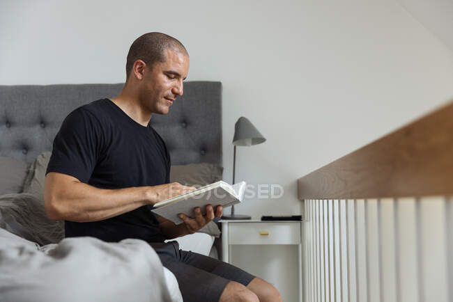 Side view of pensive male sitting on soft bed in morning and reading interesting story in book after awakening — Stock Photo