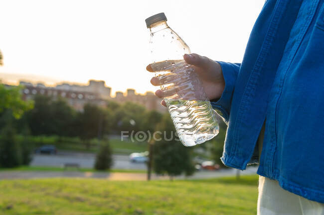 Vista lateral de la hembra sedienta irreconocible recortada bebiendo agua dulce de la botella de plástico en la ciudad en la parte posterior iluminada - foto de stock