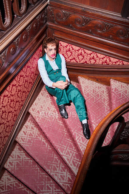Сверху эксцентричный мужской актер с размазанным макияжем сидит на лестнице во время выступления — стоковое фото