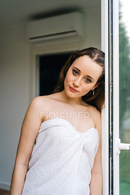 Sanftes Weibchen in weißes Handtuch gehüllt, mit nassen Haaren nach dem Duschen in der Nähe der Terrassentür und Blick in die Kamera — Stockfoto