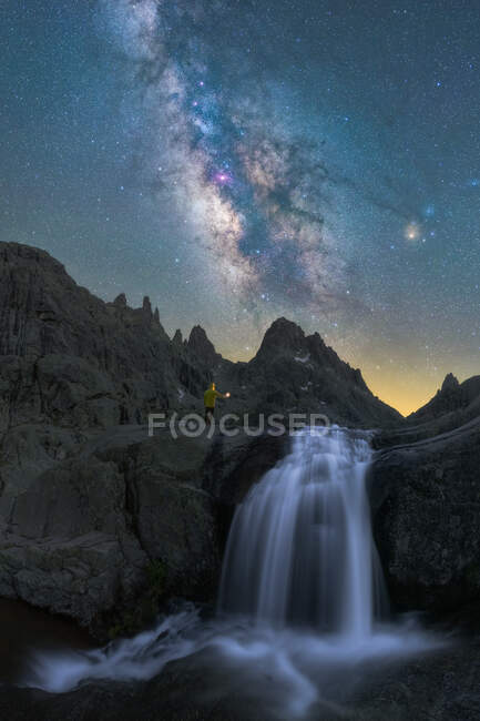 Anonymous traveler with torch contemplating waterfall streaming among rough rocky terrain under night starry sky with bright glowing Milky Way — Stock Photo