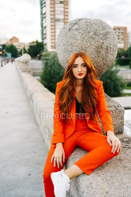 Bella femmina con lunghi capelli rossicci e in pantaloni arancione brillante seduto sul bordo sul lungomare in città guardando la fotocamera — Foto stock