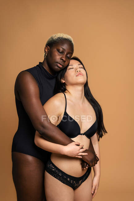 Modelli femminili multirazziali in lingerie nera che si abbracciano su sfondo beige per concetto di positività del corpo in studio con gli occhi chiusi — Foto stock