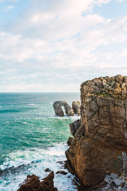 Spectaculaire paysage de rivage rocheux rugueux baigné par des vagues de mer mousseuses au soleil sous un ciel nuageux bleu à Liencres en Cantabrie en Espagne — Photo de stock