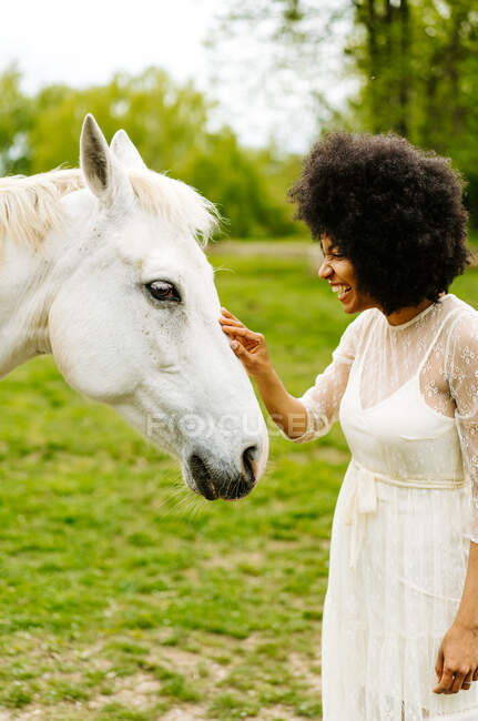 Sonriente hembra afroamericana con pelo afro rizado y vestido blanco acariciando caballo gris juntos en prado en el campo - foto de stock