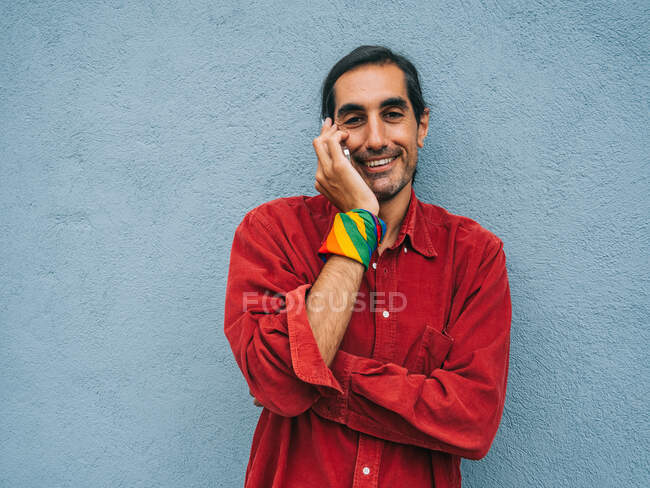 Очарованный этнический гей с радужной банданой под рукой, разговаривающий по мобильному телефону, стоя рядом с серой стеной в городе и глядя в камеру — стоковое фото