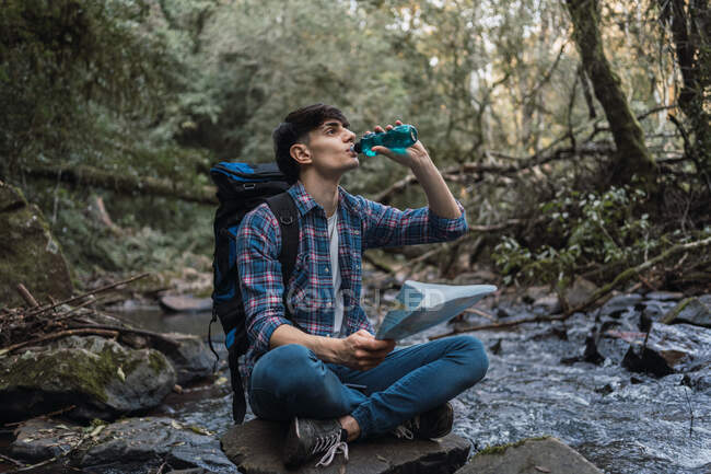 Escursionista maschio assetato che beve acqua dolce dalla bottiglia mentre è seduto sulla roccia vicino al fiume nella giungla — Foto stock