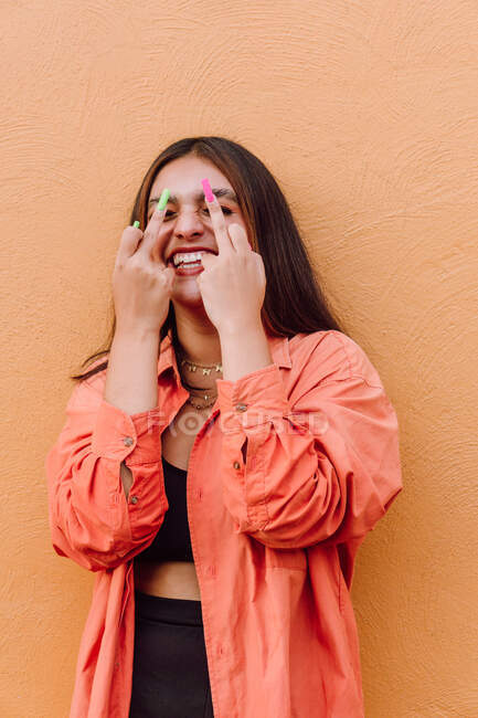 Délicieuse jeune femme élégante riant joyeusement avec les yeux fermés et montrant les doigts du milieu sur fond orange — Photo de stock