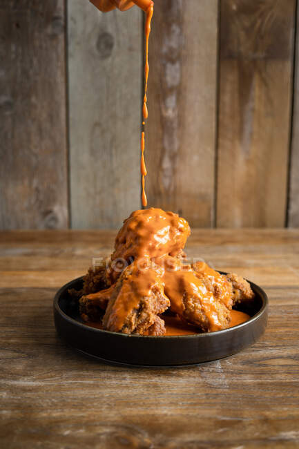 Molho saboroso derramando deliciosas asas de frango de búfalo colocadas na placa preta redonda na mesa de madeira — Fotografia de Stock
