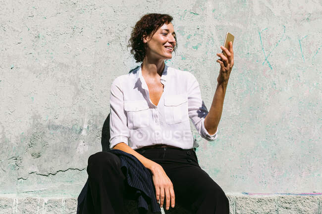 Delighted female in white formal shirt and pants sitting on concrete border and having video call — Stock Photo