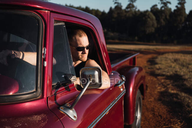 Selbstbewusster männlicher Fahrer mit Sonnenbrille sitzt in rotem Retro-Pickup, der im Sommer auf sandiger Straße im Grünen geparkt ist — Stockfoto