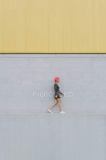 Vista lateral de la joven hembra con el pelo rosa brillante en traje elegante caminando en el borde en la pared gris en la calle - foto de stock
