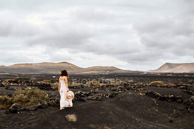 Mujer irreconocible en vestido blanco llevando sombrero y caminando sobre suelo seco cerca de arbustos en día nublado en valle sin agua en Fuerteventura, España - foto de stock