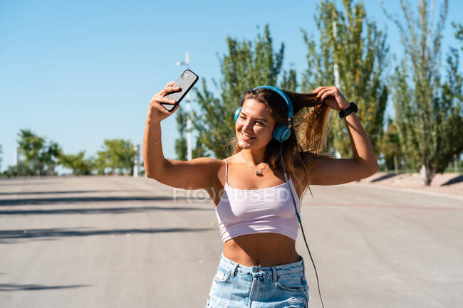 Positive female skater in rollerblades and headphones taking self shot on mobile phone on sunny day in summer in city — Stock Photo