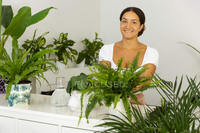 Vista lateral de la alegre hembra étnica tomando fotos de la planta en maceta en el teléfono celular en casa - foto de stock