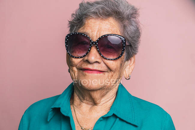 Счастливая современная женщина в седых волосах и в модных солнцезащитных очках на розовом фоне в студии и глядя в камеру — стоковое фото