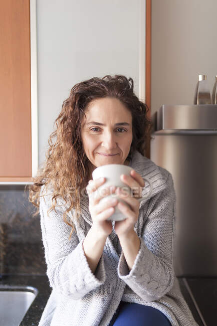 Mulher com cabelo encaracolado sentado na cozinha tomando uma infusão — Fotografia de Stock