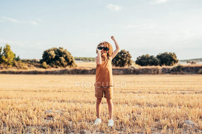Приємна етнічна дитина з африканською зачіскою дивиться через бінокль і святкує перемогу з піднятою зброєю, стоячи в сушеному одязі влітку в сонячний день і отримуючи задоволення — стокове фото