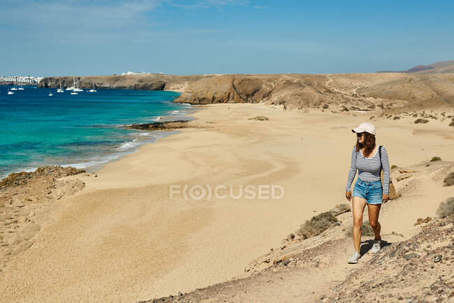 Femme en vêtements décontractés regardant loin et admirant la mer turquoise tout en marchant sur la côte sablonneuse pendant les vacances d'été Fuerteventura, Espagne — Photo de stock