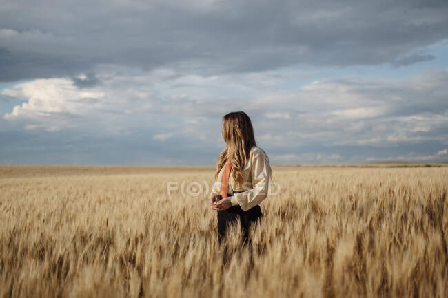 Giovane femmina con i capelli ondulati guardando lontano nel campo di campagna sotto cielo nuvoloso su sfondo sfocato — Foto stock