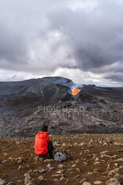 Rückansicht eines unkenntlichen Reisenden, der auf einem Berg sitzt und den Fagradalsfjall mit Feuer in Island betrachtet — Stockfoto