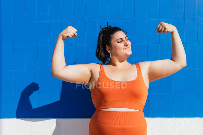 Poderoso mais atleta feminino étnico tamanho em sportswear demonstrando músculos enquanto olha para longe contra a parede de azulejos azuis na rua — Fotografia de Stock