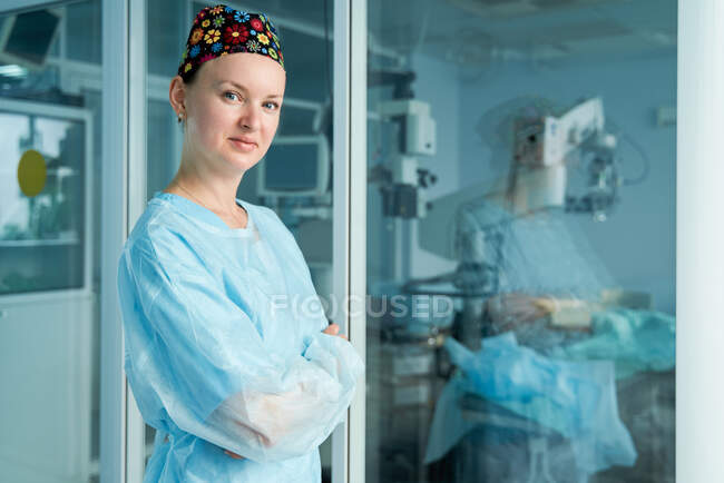 Самовпевнена жінка-лікар зі складеними руками в орнаментальній медичній шапці дивлячись на камеру проти скляної стіни в лікарні. — стокове фото