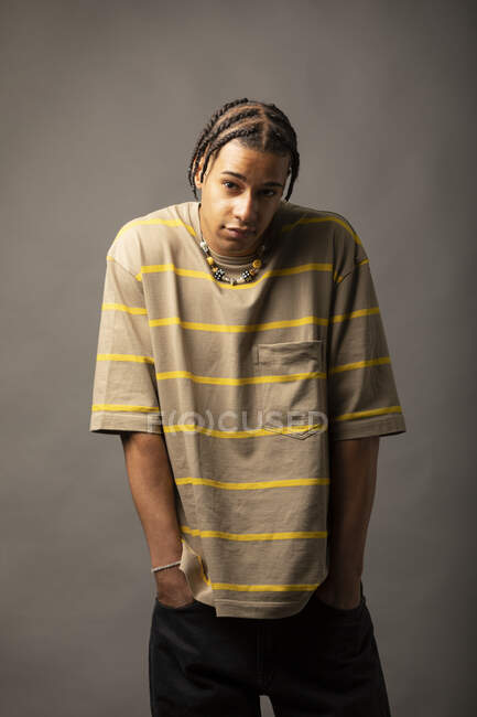 Jeune modèle masculin afro-américain avec des cheveux tressés habillés en chemise à rayures surdimensionnées et un collier regardant la caméra sur fond gris — Photo de stock