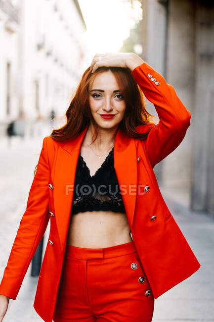 Elegante femmina con i capelli rossicci e in abito arancione vivace passeggiando per la strada della città guardando la fotocamera — Foto stock