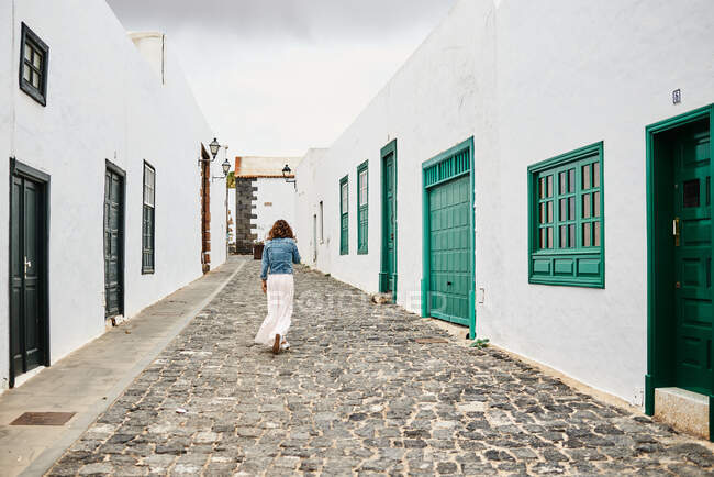 Задний вид анонимной женщины в повседневной одежде, идущей по каменному тротуару возле белых домов на улице города Фуэртевентура, Испания — стоковое фото