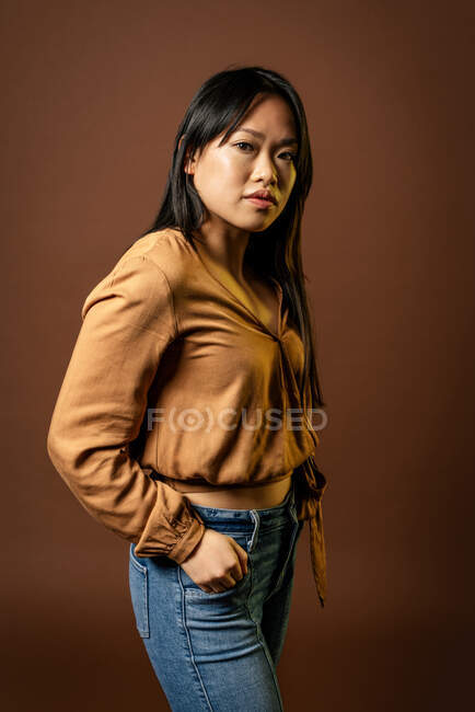 Vista lateral da mulher asiática em roupas da moda olhando para a câmera no fundo marrom em estúdio — Fotografia de Stock