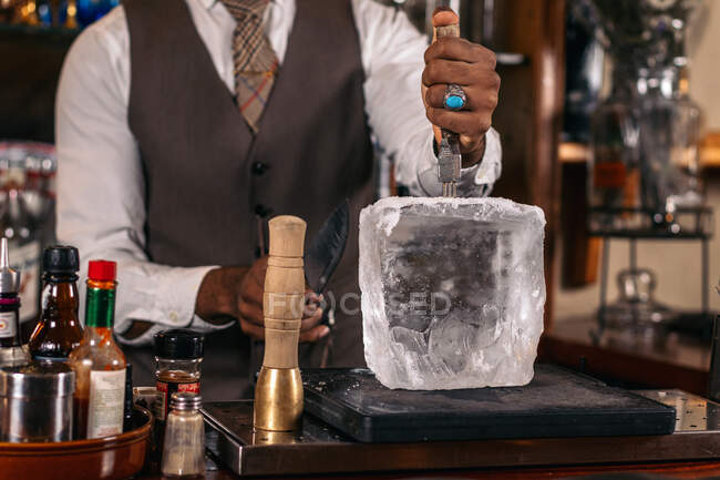 Crop unrecognizable stylish ethnic male barkeeper in ring crushing ice cube with tool at work in bar — Stock Photo
