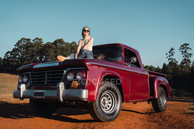 Low angle of delighted cool female sitting on hood of red vintage pickup car parked in countryside in summer evening — Stock Photo