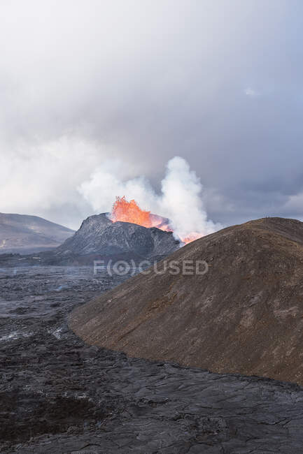 Живописный вид на Fagradalsfjall с быстрым огнем и лаву под рассеивающимся дымом в горах с облаками в Исландии — стоковое фото