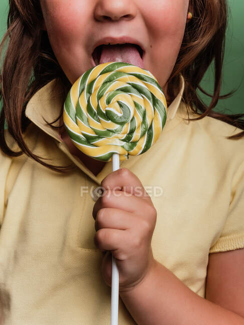 Criança anônima estendendo a mão com pirulito doce redemoinho em direção à câmera no fundo verde no estúdio — Fotografia de Stock