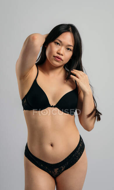Mulher asiática confiante em lingerie preta em pé sobre fundo cinza em estúdio e olhando para a câmera — Fotografia de Stock