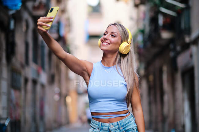 Беззаботная женщина в жёлтых наушниках слушает музыку и делает автопортрет на смартфоне, развлекаясь летом на городской улице. — стоковое фото