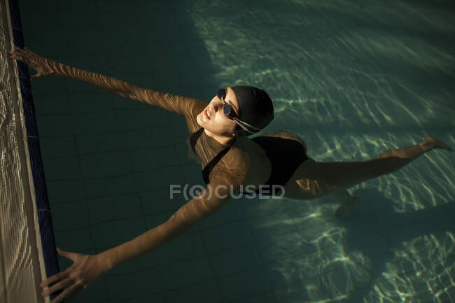 Giovane bella donna sul marciapiede della piscina coperta, indossando costume da bagno nero, galleggiante in acqua — Foto stock