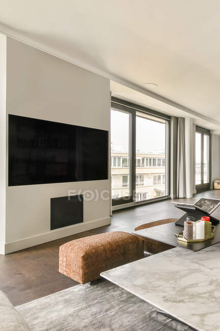 Дизайн інтер'єру стильної просторої вітальні з великим телевізором, що висить на білій стіні перед столом і м'якими кушетками, прикрашеними горщиками в сучасній розкішній квартирі — стокове фото