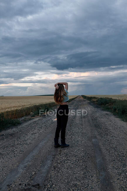 Stylish young woman touching long hair on roadway under cloudy sky in twilight — Stock Photo