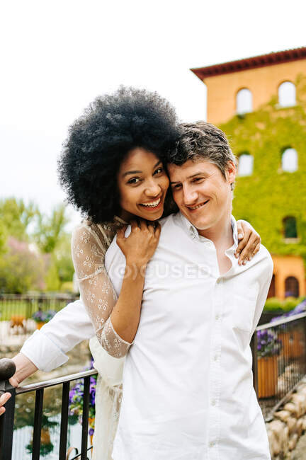 Loving black woman with Afro hairstyle hugging smiling man from behind while standing on bridge in park in summer and enjoying weekend together — Stock Photo