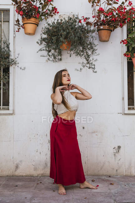 Mulher despreocupada com lábios vermelhos e roupas de verão em pé descalço no pátio da casa perto da parede com flores em vasos pendurados e pescoço tocante com olhos fechados — Fotografia de Stock