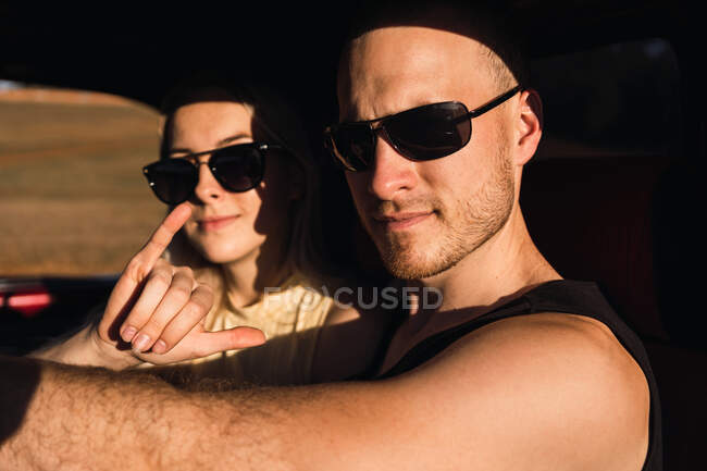 Copain cool montrant geste shaka tandis que assis dans la voiture avec petite amie cool dans des lunettes de soleil le jour ensoleillé — Photo de stock